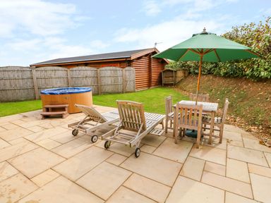 An outdoor patio with wooden chaise lounges a table with chairs and a green umbrella next to a hot tub and a wooden fence in the background at Cedar Lodge in Greetham Rutland