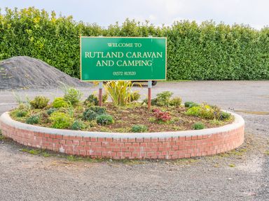 A green welcome sign for Rutland Caravan and Camping on a circular brick planter with plants and gravel ground