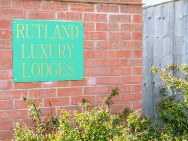 A green sign with yellow text reading Rutland Luxury Lodges on a brick wall with green shrubs and a wooden fence