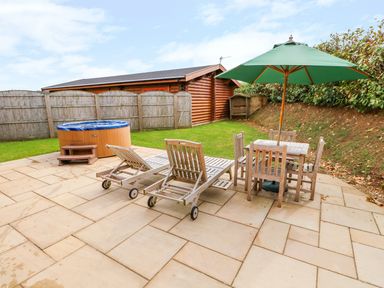 A backyard patio with two wooden lounge chairs a wooden table with chairs and a green umbrella and a hot tub near a wooden fence and a log cabin at Beech Lodge in Greetham Rutland