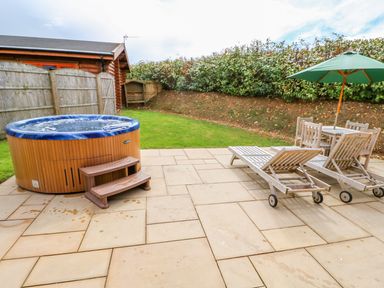 An outdoor patio with a hot tub, two wooden lounge chairs, a table with four chairs, and a green umbrella at Beech Lodge in Greetham Rutland
