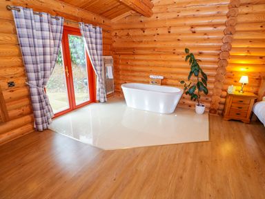 A room with wooden walls a bathtub by a glass door with curtains and a bedside table with a lamp at Beech Lodge in Greetham Rutland