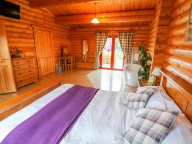 A bedroom with a wooden bed, purple blanket, plaid pillows, a dresser, a desk with a mirror, a bathtub, and curtains on glass doors at Beech Lodge in Greetham Rutland