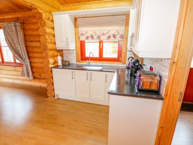 A kitchen with white cabinets black countertop a toaster kettle and coffee machine a window with floral blinds and wooden log walls at Beech Lodge in Greetham Rutland