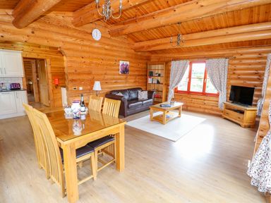 A log cabin living room with a wooden dining table and chairs a brown leather sofa a coffee table bookshelf television on a stand and floral curtains at Beech Lodge Greetham Rutland