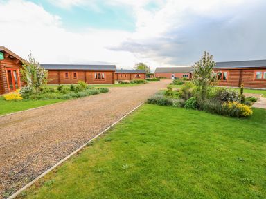 A gravel driveway with grass and shrubs on either side with wooden cabins and a brick building in the background at Beech Lodge in Greetham Rutland