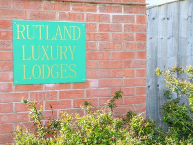 A green sign reading Rutland Luxury Lodges on a red brick wall with green bushes and a wooden fence