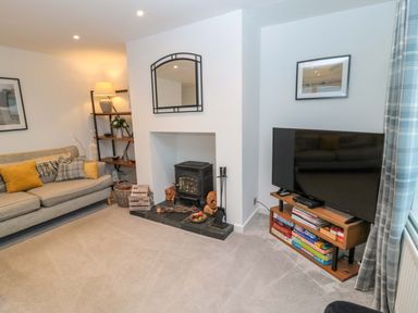 A living room with a sofa cushions a fireplace with wood a television on a stand with board games and a shelving unit with a lamp at Wynding Apartment in Amble-By-The-Sea