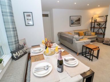 A dining table set for four next to a seating bench with cushions beside a living room with two sofas and a bookshelf at Wynding Apartment in Amble-By-The-Sea