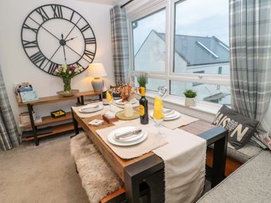 A dining area with a wooden table set for four and a large wall clock at Wynding Apartment in Amble-By-The-Sea