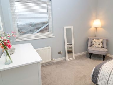 A bedroom corner with a chair and floor lamp next to a mirror and white dresser with flowers at Wynding Apartment in Amble-By-The-Sea