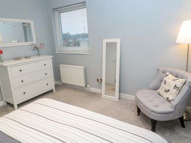 A bedroom with a bed, white dresser, mirror, gray chair with a dog print pillow and a floor lamp at Wynding Apartment in Amble-By-The-Sea