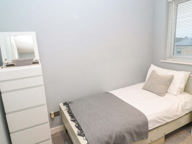 A small bedroom with a single bed a gray blanket a white pillow a bedside drawer with a mirror and a window with blinds at Wynding Apartment in Amble-By-The-Sea