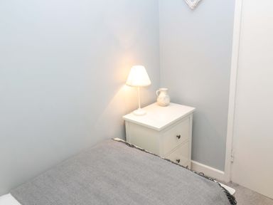 A bedside table with a lamp and a vase next to a bed with a gray blanket at Wynding Apartment in Amble-By-The-Sea
