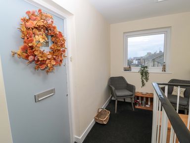 A hallway with a door decorated with a wreath two chairs and a window showing rooftops at Wynding Apartment in Amble-By-The-Sea