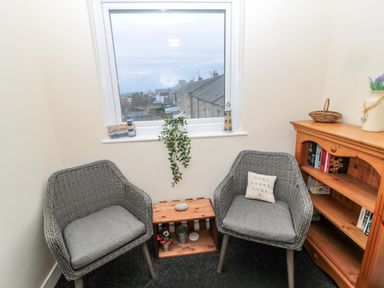 A small seating area with two grey wicker chairs featuring grey cushions and a wooden bookshelf near a window at Wynding Apartment in Amble-By-The-Sea