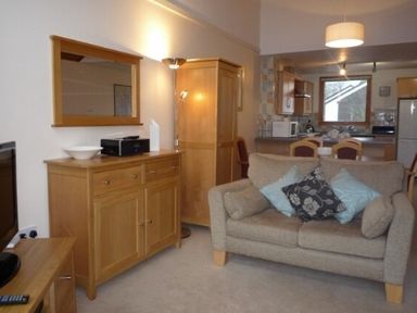A living area with a sofa and cushions a wooden cabinet with a mirror and a kitchen in the background at 13 Keswick Bridge in Keswick