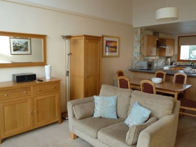 A living area with a beige sofa and light blue cushions a wooden cabinet with a white vase and a stereo a floor lamp a dining table with chairs and a kitchen with wooden cabinets at 16 Keswick Bridge in Keswick