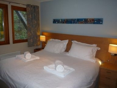 A bedroom with a double bed white bedding rolled towels bedside tables with lamps and windows with curtains at 16 Keswick Bridge in Keswick