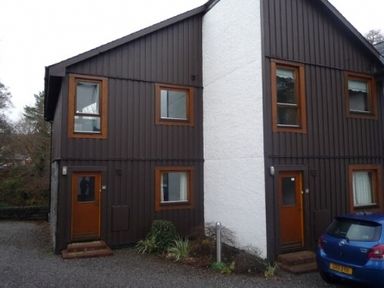 Two connected brown wooden houses with orange doors and windows and a blue car parked outside at 16 Keswick Bridge in Keswick