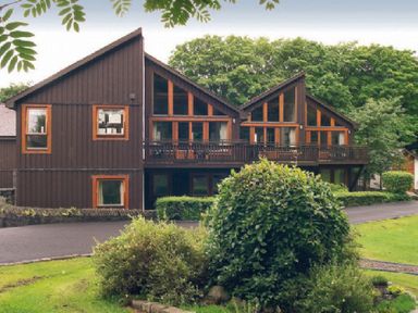 A brown wooden building with large windows and balcony surrounded by green bushes and grass at 20 Keswick Bridge in Keswick