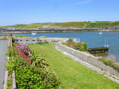 A garden with flowers and a lawn next to a body of water with boats and a pier at Angorfa in Cemaes Bay