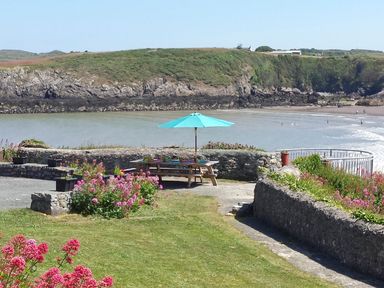 A picnic table with an umbrella near a stone wall overlooking a beach and rocky cliffs at Angorfa in Cemaes Bay