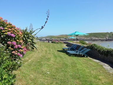 A grassy garden area with lounge chairs and umbrellas next to a stone wall by the sea at Angorfa in Cemaes Bay