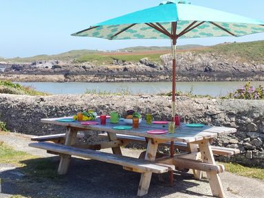 An outdoor picnic table with colorful plates cups and an umbrella by a stone wall and water at Angorfa in Cemaes Bay