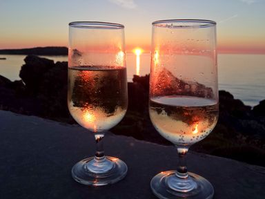 Two glasses with a light beverage on a surface with a sunset over water and rocks in the background at Angorfa in Cemaes Bay