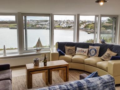 A living room with a curved cream sofa with cushions and a wooden coffee table with wine and glasses by large windows overlooking water and houses at Angorfa in Cemaes Bay