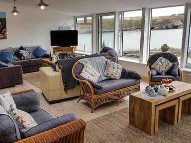 A living room with wicker chairs sofas a wooden coffee table and large windows overlooking water at Angorfa in Cemaes Bay