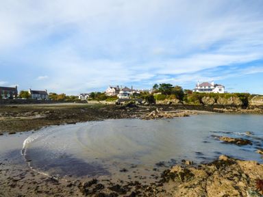 A rocky shoreline with houses on a cliff under a blue sky at Angorfa in Cemaes Bay