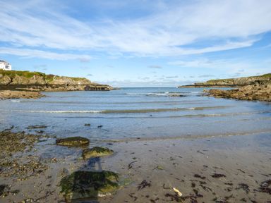 A sandy beach with rocks and seaweed leading to calm ocean water with rocky cliffs on both sides at Angorfa in Cemaes Bay