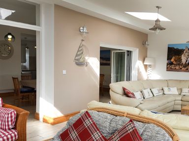 A living room with a beige sectional sofa with cushions and a wicker chair with red plaid pillows at Angorfa in Cemaes Bay