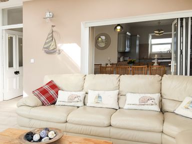 A beige leather sofa with decorative cushions in a living room with a dining area and kitchen visible through an open partition wall at Angorfa in Cemaes Bay