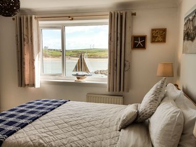 A bedroom with a bed near a window with curtains and a model sailboat on the windowsill at Angorfa in Cemaes Bay