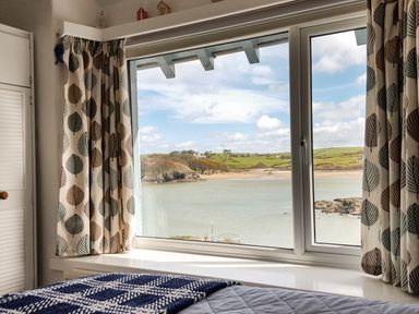 A bedroom window with curtains overlooking a bay and green hills at Angorfa in Cemaes Bay