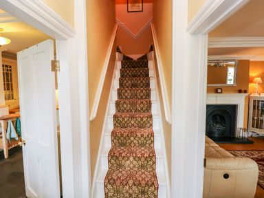 A staircase with patterned carpet runner between two open doors showing a kitchen on the left and living room with fireplace on the right at Castle School House in Kidwelly
