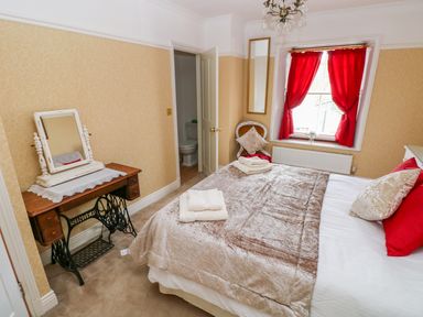 A bedroom with a bed, red curtains, a vintage sewing machine table with a mirror, and a chair by the window at Castle School House in Kidwelly