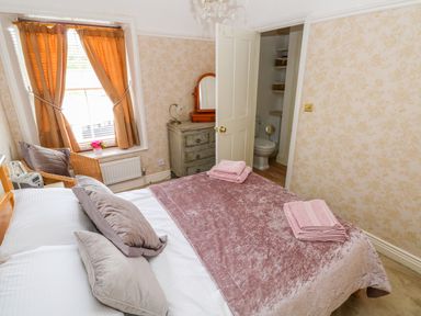A bedroom with a bed with pillows and pink towels a wicker chair near a window and a dresser next to an open door showing a toilet at Castle School House in Kidwelly