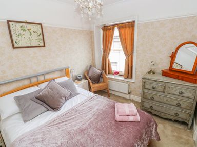 A bedroom with a made bed with pillows and folded towels a wicker chair by a window and a dresser with mirror at Castle School House in Kidwelly