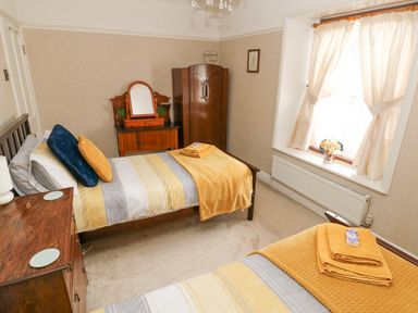A bedroom with two beds yellow and gray bedding and towels a wooden wardrobe and a window with curtains at Castle School House in Kidwelly