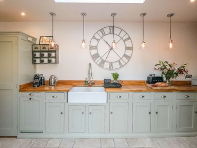 A kitchen with light gray cabinets wooden countertop a white farmhouse sink hanging lights large wall clock and flowers at Harriets Cottage in Briggswath