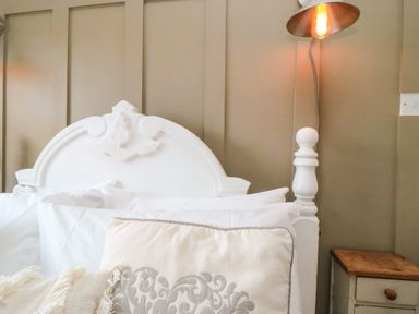 A white headboard with pillows and a beige decorative cushion next to a wooden bedside table with a lamp on the wall at Harriets Cottage in Briggswath