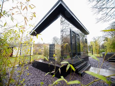 A small modern building with mirrored walls and black steps surrounded by plants and trees at Reflection in Longtown