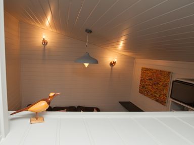 A room with white paneled walls and ceiling wall lights a hanging ceiling lamp a wooden bird sculpture a colorful painting a microwave and a dark shelf at Reflection in Longtown