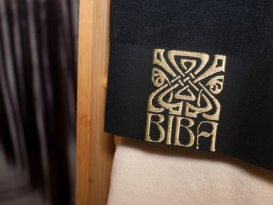 A close up of black and beige towels hanging on a wooden rack with an embroidered gold design and text saying BIBA