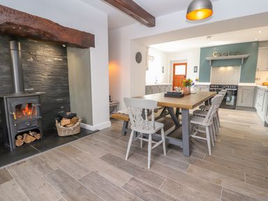 A kitchen dining area with a wooden dining table and gray chairs a wood burning stove with fire and basket of logs in a stone alcove at Llys Alaw in Llangernyw