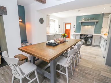 A kitchen with a wooden dining table and white chairs next to a stove under a shelf with the word Croeso at Llys Alaw in Llangernyw